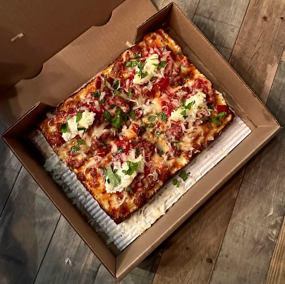 A rectangular Detroit-style pizza with thick, crispy caramelized edges, topped with red sauce, melted cheese, dollops of white ricotta, and fresh green basil, served in a cardboard delivery box.