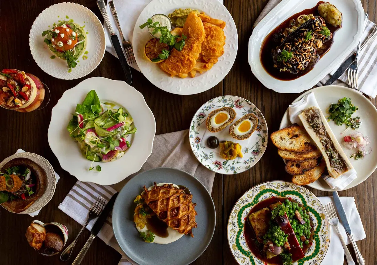 A flat lay view of a dark wooden table filled with a variety of gourmet dishes, including fish and chips, a scotch egg, bone marrow with toast, a fresh green salad, and other beautifully plated entrees.