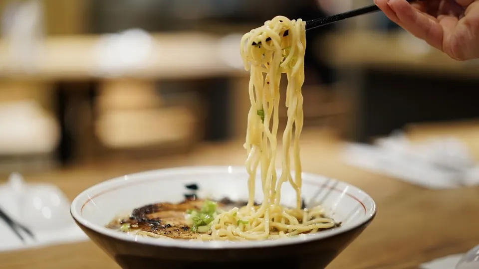 A close-up of thick ramen noodles being lifted from a steaming bowl of ramen with chopsticks, showing grilled pork chashu and green onions in a rich broth.