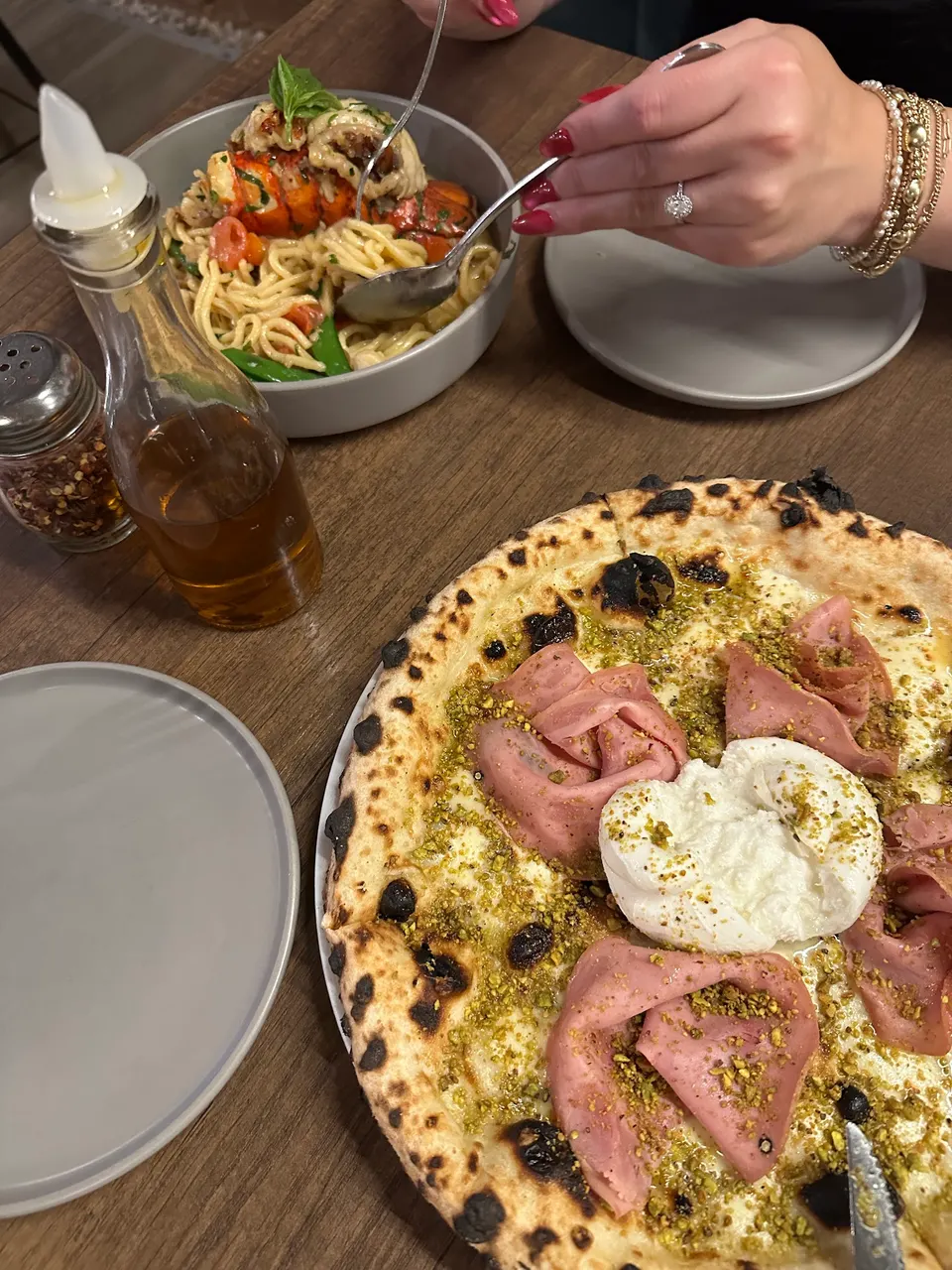 A table setting at Pizza e Pazzi featuring a gourmet pizza topped with mortadella, burrata, and pistachios, alongside a bowl of lobster pasta being served.