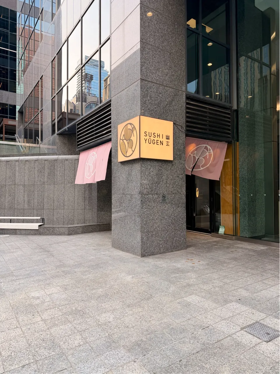 The exterior entrance of Sushi Yugen, featuring a modern grey stone pillar with a glowing square corner sign, pink fabric noren curtains, and a paved outdoor plaza.
