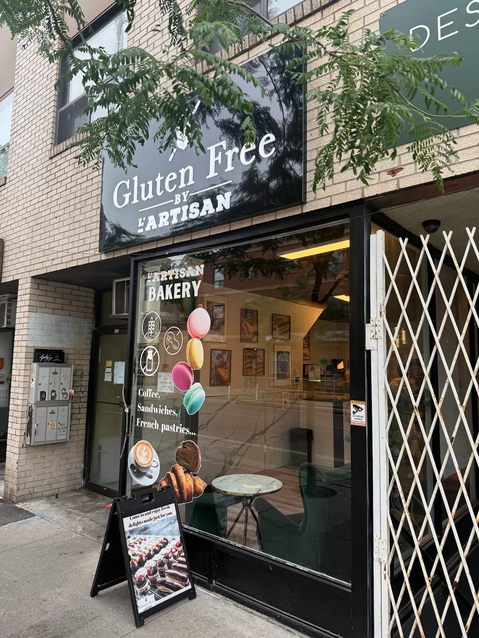 The storefront of Gluten Free by L'Artisan, featuring a black sign with white text, a large window displaying bakery offerings, and a sidewalk sandwich board sign.