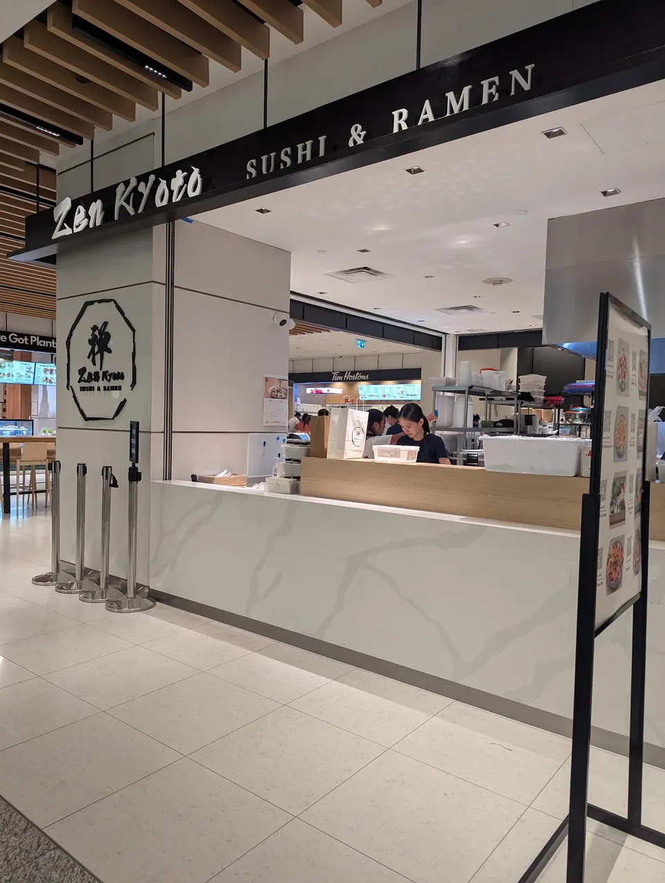 The storefront of Zen Kyoto Sushi & Ramen in a modern food court, featuring a white marble counter, black signage, and staff preparing food behind the counter.