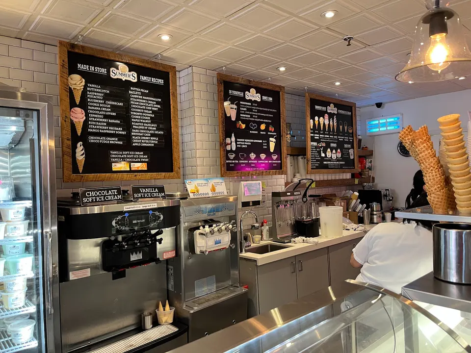 The counter area of Summer's Ice Cream shop, featuring soft serve machines, menu boards listing flavors, and stacks of waffle cones on the counter.