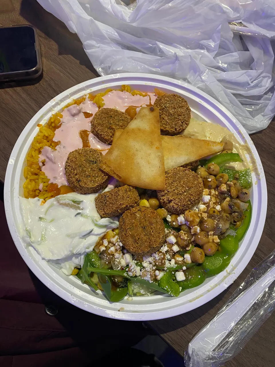 A Greek street food bowl filled with yellow rice, falafel, tzatziki, hummus, chickpeas, greens, feta cheese, and pita chips.