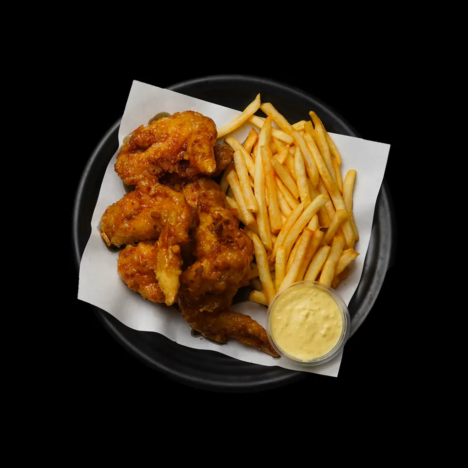 A top-down view of a black plate containing crispy glazed chicken wings, a generous portion of golden french fries, and a small cup of creamy dipping sauce on white parchment paper.