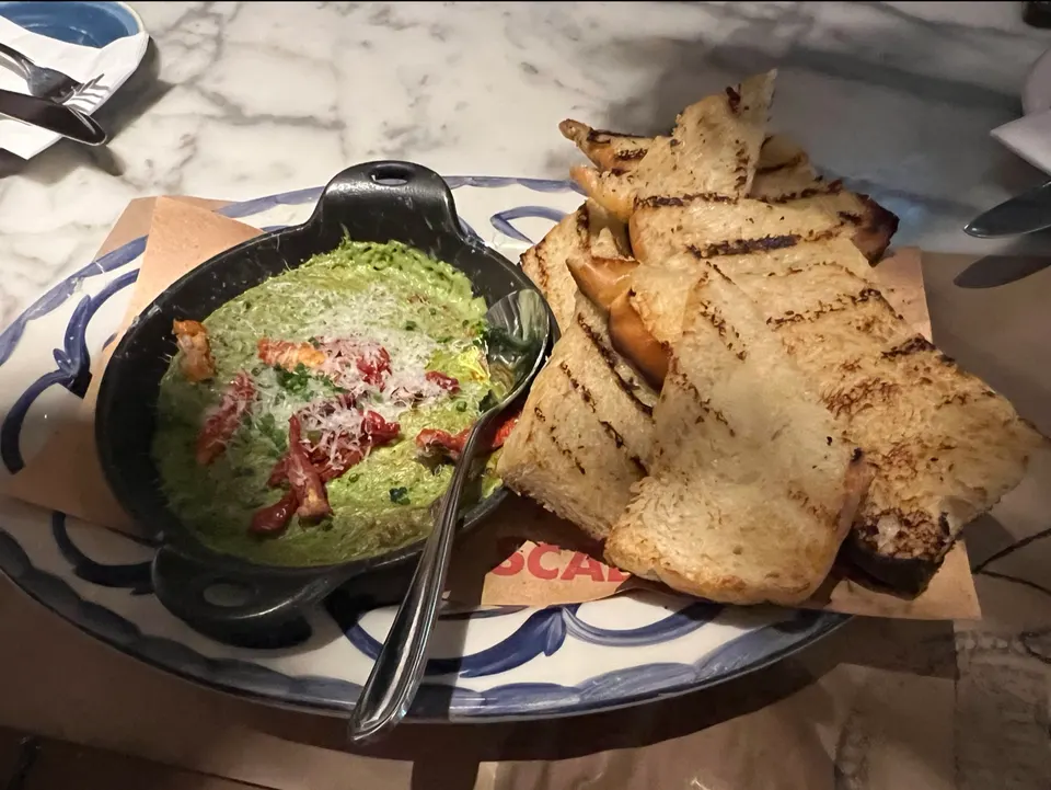 A black skillet filled with a green pesto dip topped with sun-dried tomatoes and grated cheese, served on a platter alongside several slices of grilled bread.