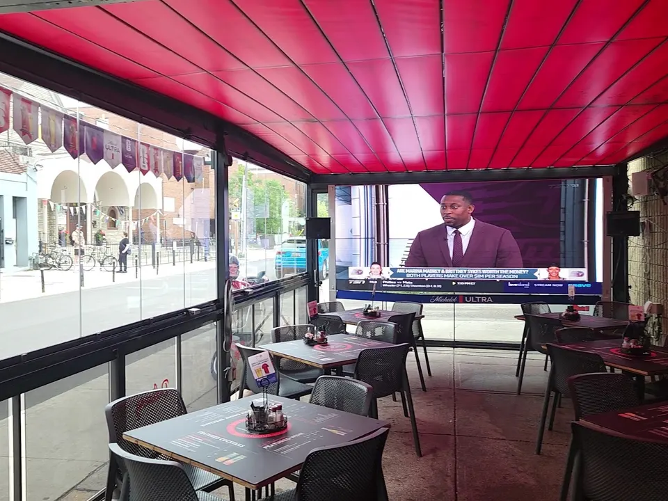 An outdoor covered patio dining area at Cafe Diplomatico with black tables and chairs, a bright red retractable awning, and a large television screen showing a sports broadcast.