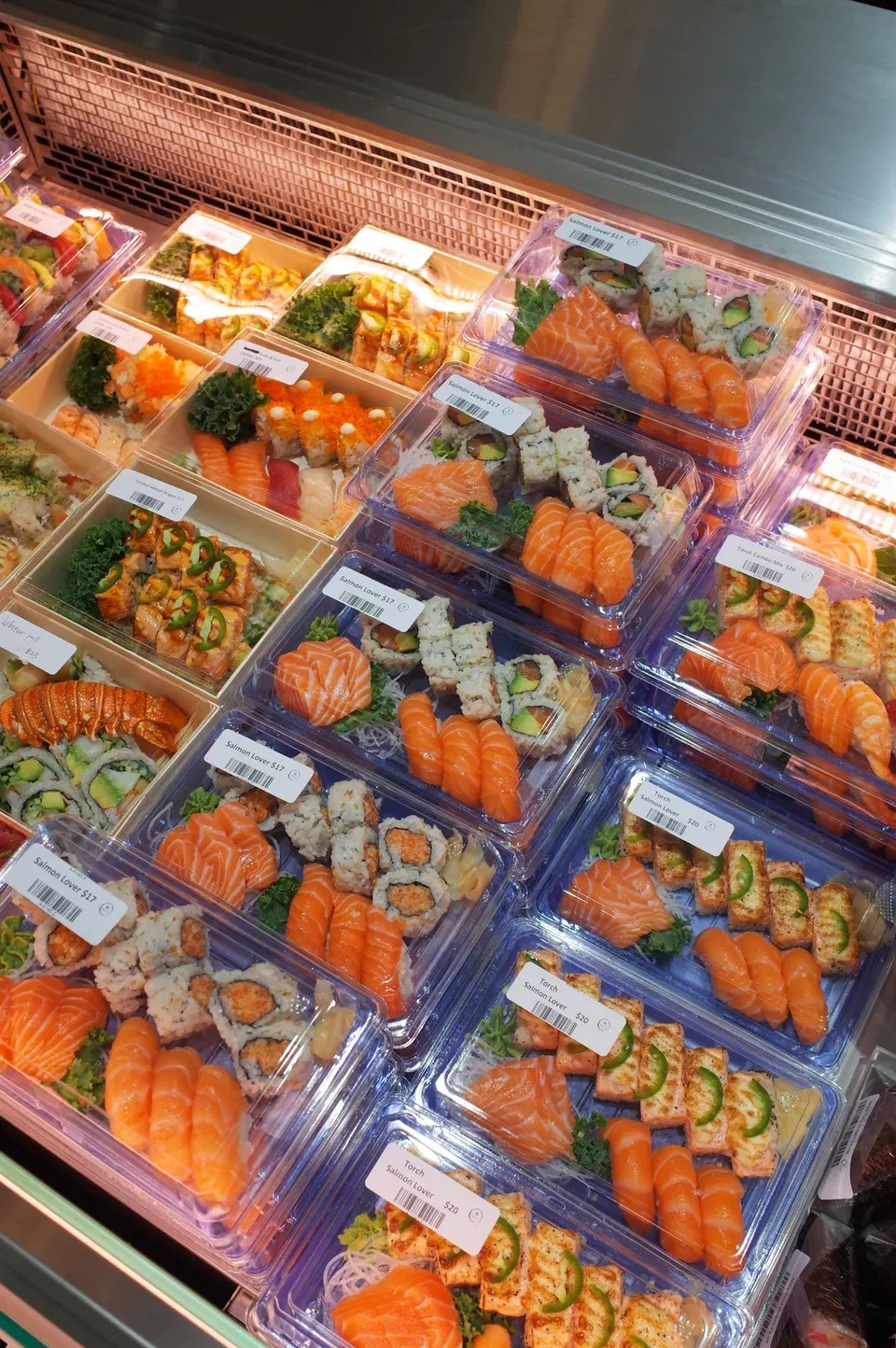 A display case filled with pre-packaged sushi trays, featuring salmon nigiri, maki rolls, and seared salmon with jalapeño toppings, each labeled with price tags.