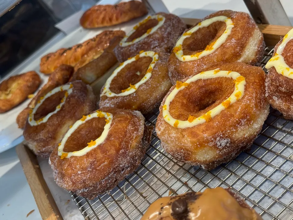 A close-up shot of several sugar-dusted cronuts topped with white icing rings and orange zest, arranged on a wire cooling rack inside a bakery display case.