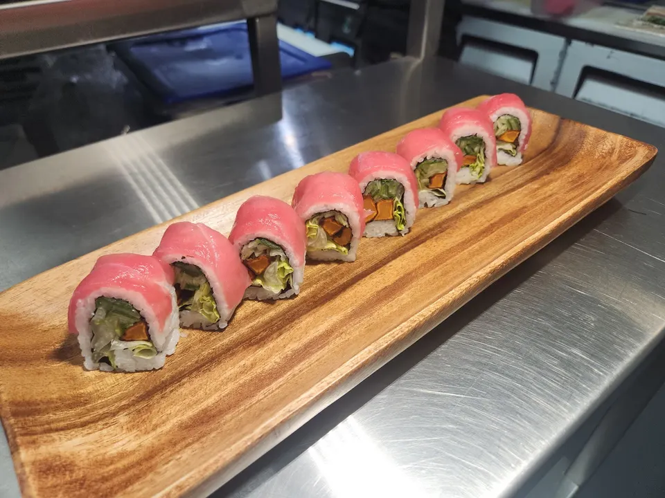 Eight pieces of sushi roll topped with bright pink tuna, filled with greens and sweet potato, arranged diagonally on a long wooden serving board on a stainless steel counter.
