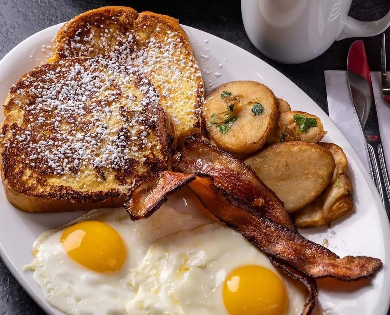 A breakfast plate featuring two sunny-side-up eggs, thick-cut crispy bacon, roasted breakfast potatoes, and two slices of powdered sugar-dusted French toast.