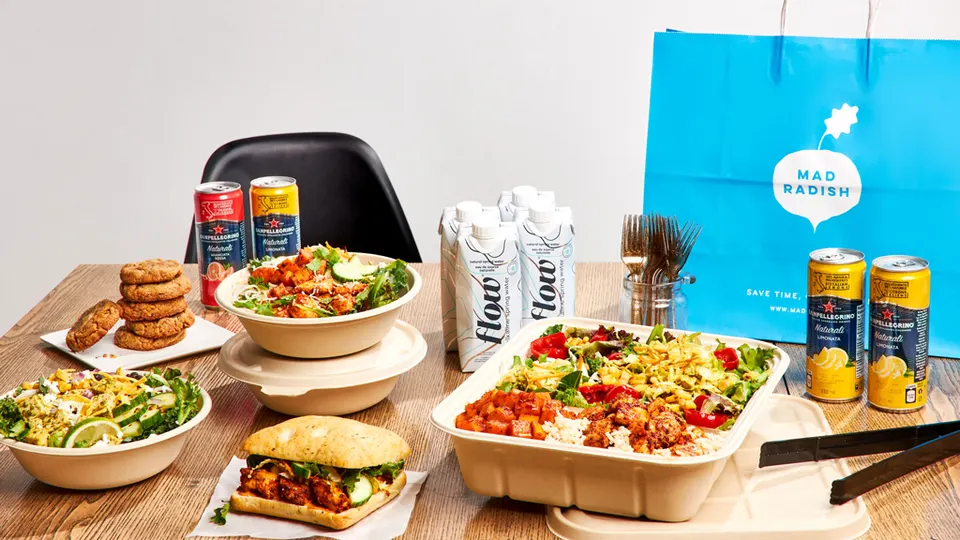 A wooden table spread with healthy catering options from Mad Radish, including salad bowls, a chicken sandwich, a large catering platter, canned beverages, and boxed water next to a blue branded paper bag.