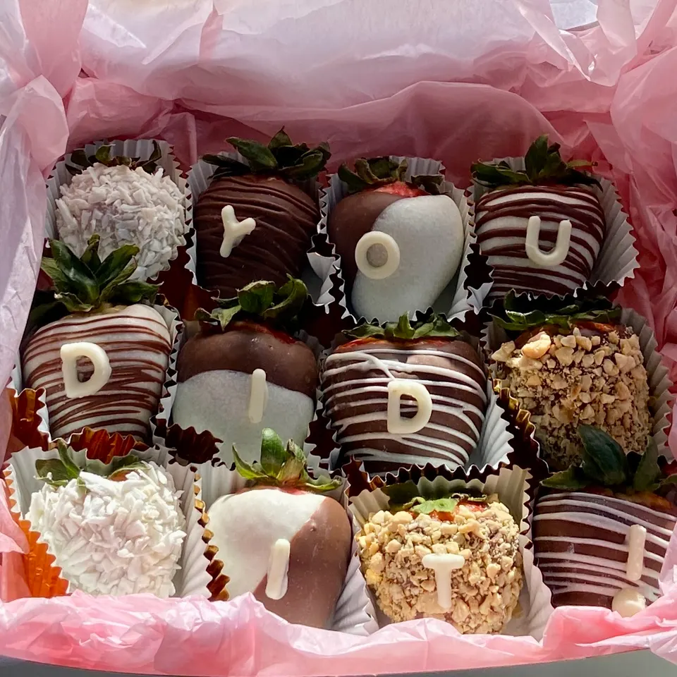 A box of twelve chocolate-covered strawberries decorated with white chocolate letters spelling out 'YOU DID IT!' alongside coconut and nut toppings.