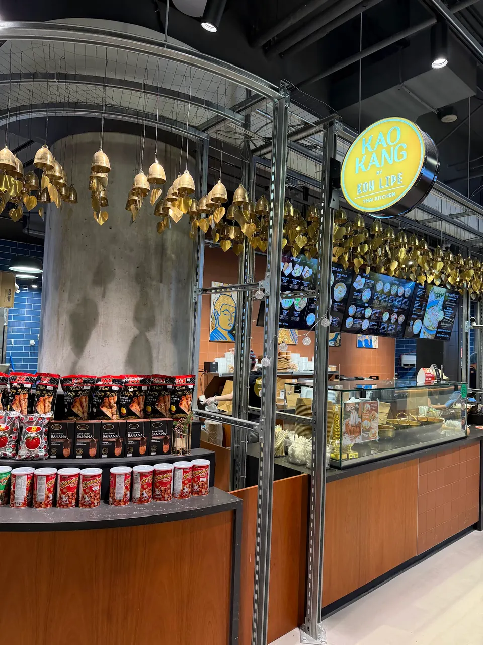 The storefront of Kao Kang by Koh Lipe, a Thai restaurant in Wellington Place, featuring a wooden counter, hanging gold bells, and a display of packaged snacks.