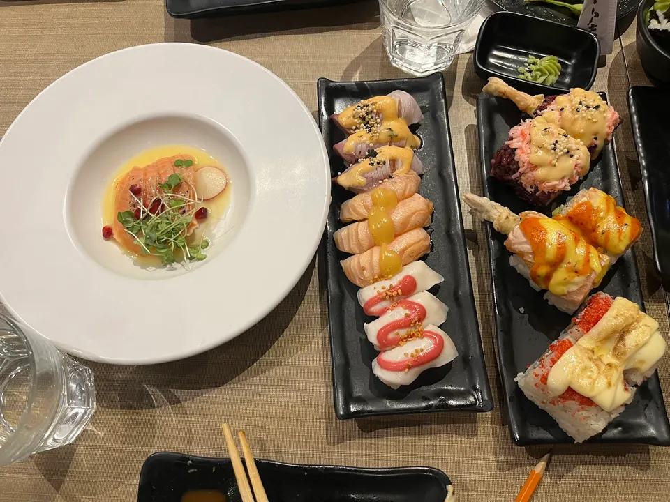 An overhead shot of a table at KAKA All You Can Eat featuring a white plate with salmon carpaccio and two black rectangular platters filled with assorted sushi rolls and nigiri.