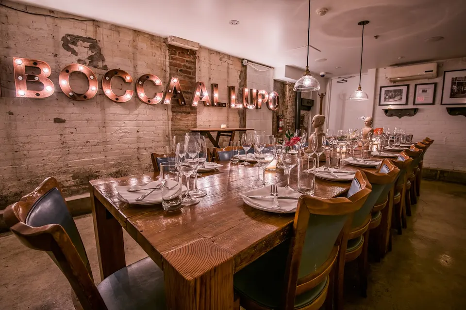 A long wooden dining table set for a group dinner with wine glasses, plates, and cutlery, positioned in front of a rustic brick wall featuring a large illuminated 'BOCCA AL LUPO' sign.