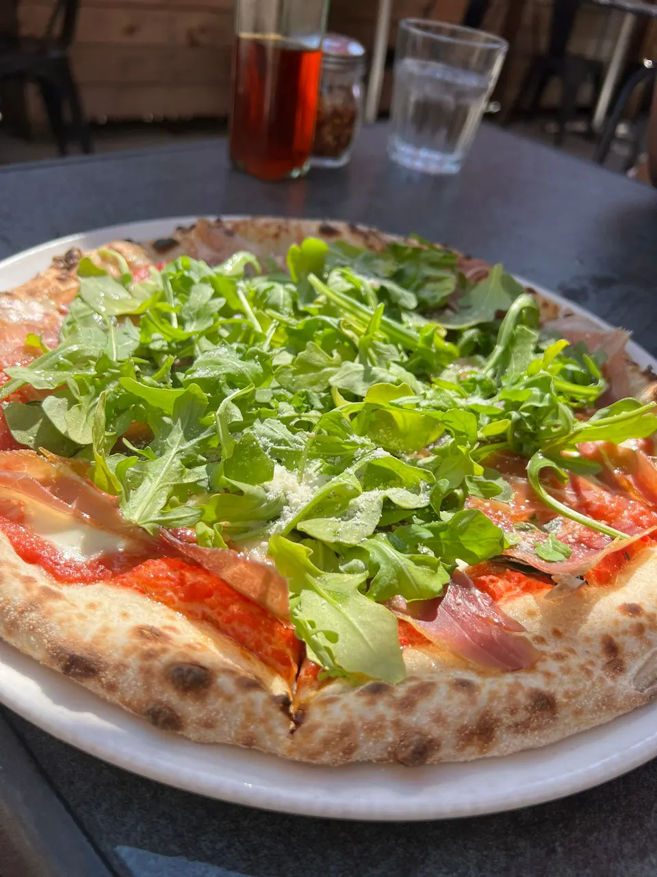 A freshly baked wood-fired pizza topped with tomato sauce, melted cheese, prosciutto slices, and a generous mound of fresh green arugula, served on a white plate on an outdoor table.