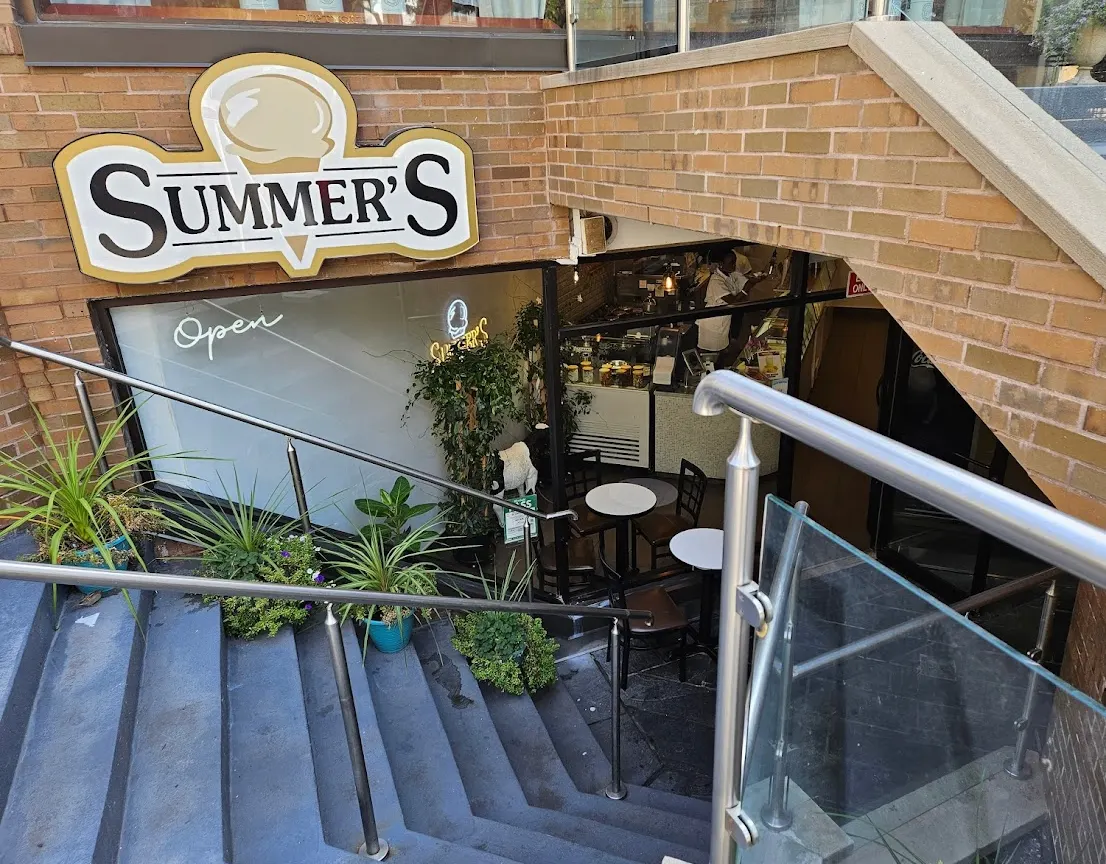 An outdoor view looking down a set of blue stairs toward the entrance of Summer's Ice Cream, featuring a large sign with an ice cream cone graphic and a small patio area with tables.