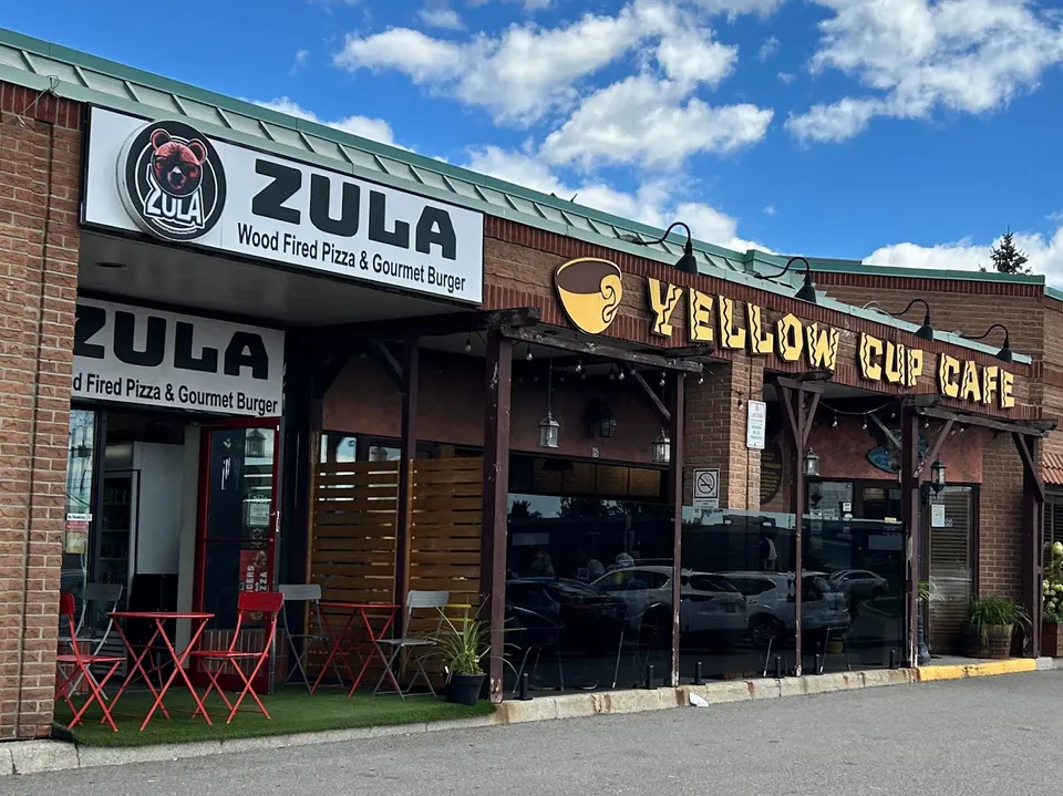 Exterior storefront of Zula Wood Fired Pizza & Gourmet Burger next to Yellow Cup Cafe, featuring outdoor seating with red and grey chairs on a small patch of artificial turf.