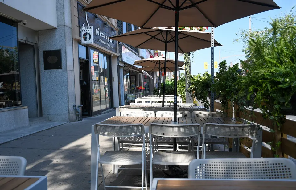 A sidewalk patio dining area with wooden tables, white metal chairs, and large brown umbrellas set up in front of Pizza e Pazzi restaurant.