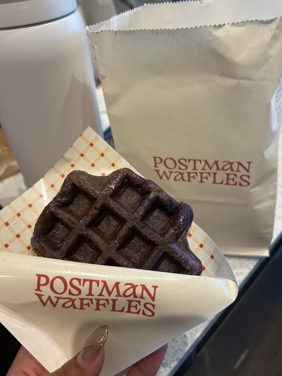 A hand holds a dark purple waffle in a paper wrapper with 'POSTMAN WAFFLES' printed on it. Another paper bag with the same branding is visible in the background, next to a white insulated bottle.