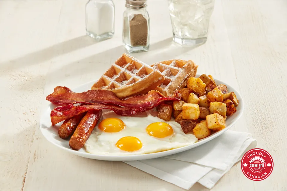 A breakfast platter featuring three sunny-side-up eggs, two sausages, strips of bacon, home fries, and two waffle quarters dusted with powdered sugar, served on a white plate with salt, pepper, and water in the background.
