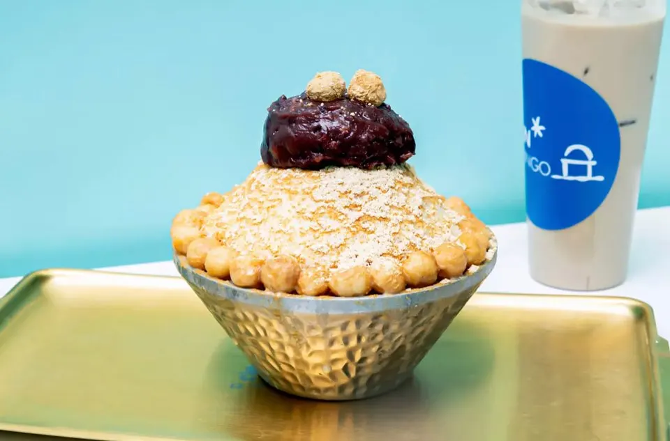 A bowl of Korean bingsu shaved ice topped with red bean paste, injeolmi powder, and rice cakes, served on a gold tray next to an iced milk tea drink.