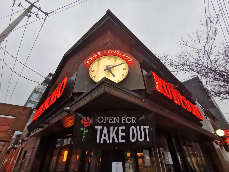 Low-angle view of a corner building exterior featuring a large clock under a neon sign reading King & Portland, with a banner below saying Open For Take Out.