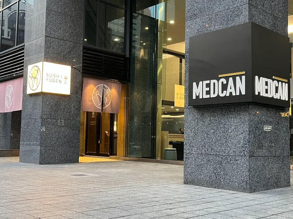 Exterior view of Sushi Yugen entrance with a glowing square sign and purple fabric noren curtains, next to a large black and white Medcan sign on a grey stone pillar.