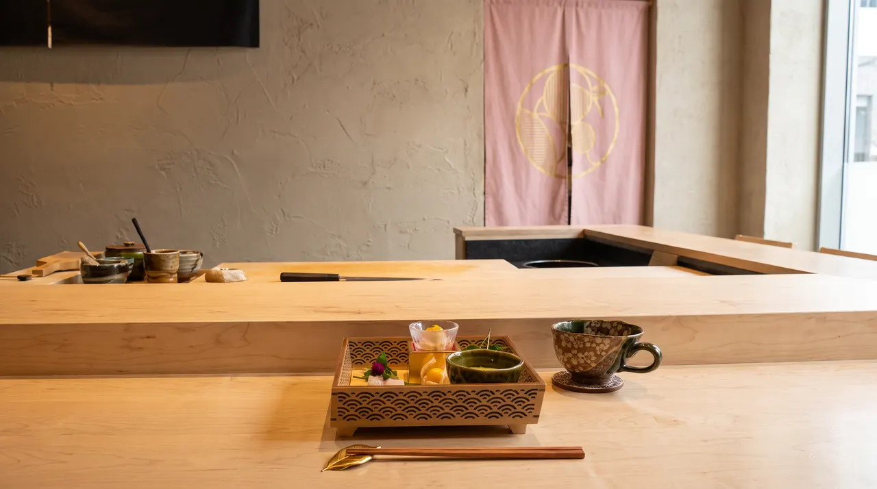 A wooden sushi counter at Sushi Yugen featuring a small decorative bento box with appetizers, a ceramic cup, and chopsticks on a gold holder.
