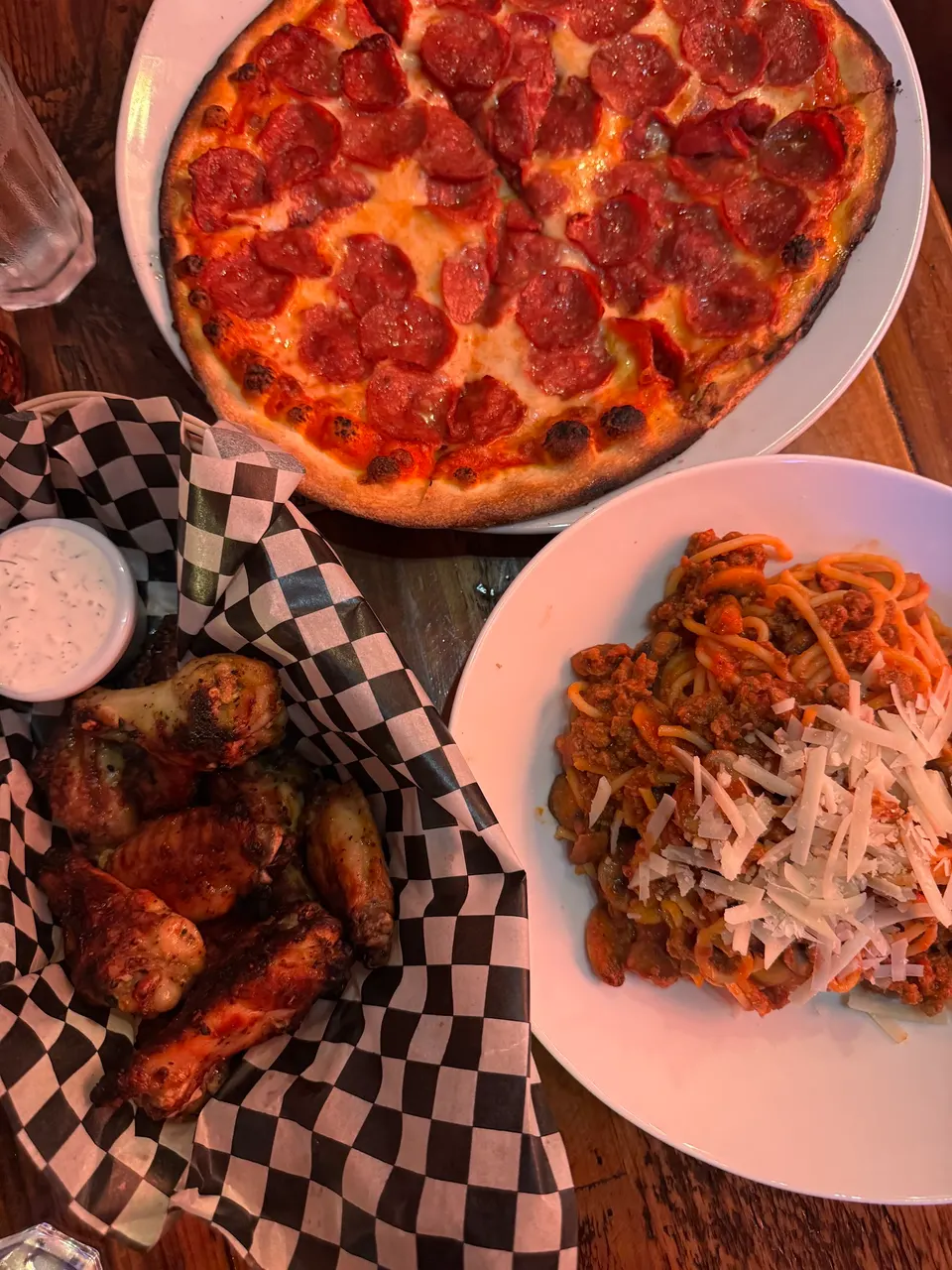 A top-down view of a table spread featuring a whole pepperoni pizza, a plate of spaghetti bolognese topped with shaved parmesan cheese, and a basket of roasted chicken wings with a side of dipping sauce.