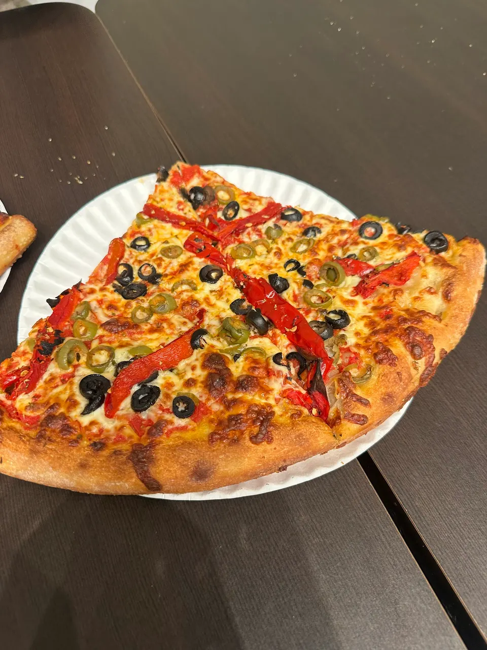 Two large slices of vegetarian pizza topped with black olives, green olives, and roasted red peppers on a white paper plate, resting on a dark wooden table.