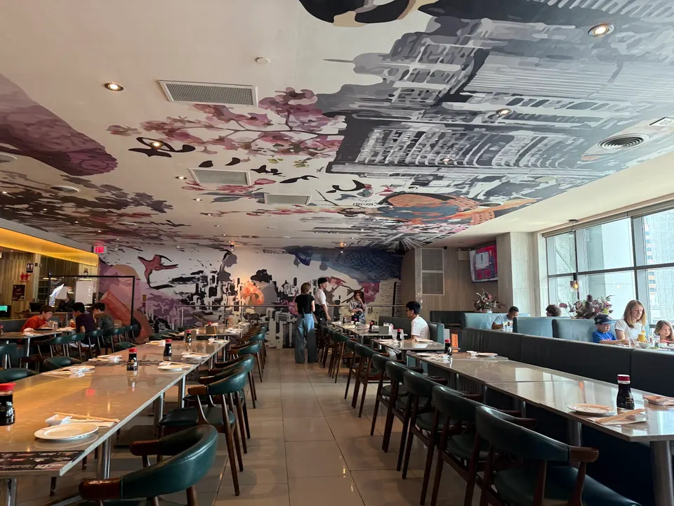 Spacious dining room at Spring Sushi Toronto with long wooden tables, dark green chairs, and a large, colorful mural covering the ceiling and back wall.