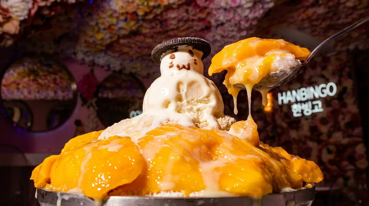 A large bowl of mango bingsu topped with fresh mango chunks, condensed milk, and a cute snowman made of ice cream wearing an Oreo cookie hat, with a spoonful being lifted.