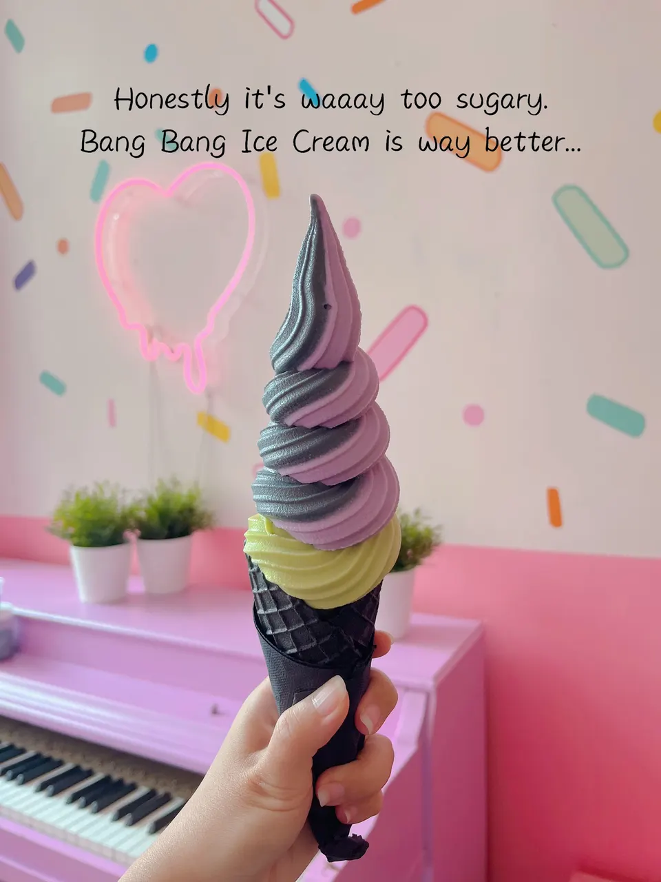 A hand holding a tall soft-serve ice cream cone with layers of grey, purple, and green swirls in a black waffle cone, set against a pink piano and a decorated wall.