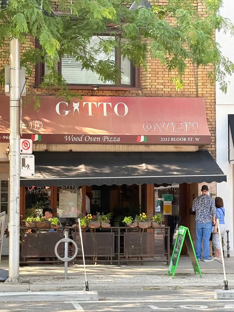 Exterior storefront of GATTO Wood Oven Pizza with a red awning, black patio canopy, outdoor seating area with flower boxes, and customers standing near the entrance.