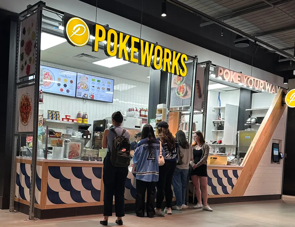 The front counter of a Pokeworks restaurant with its prominent yellow and black sign. Several customers are standing in line, facing the counter where menu screens display food options. The counter features a blue and white scalloped design.
