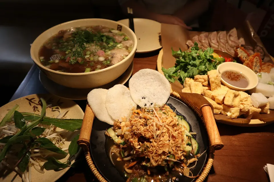 A table spread of Vietnamese dishes including a large bowl of beef pho, a plate of fresh herbs and bean sprouts, a salad topped with fried shallots and rice crackers, and a platter of fried tofu, herbs, and sliced meats.