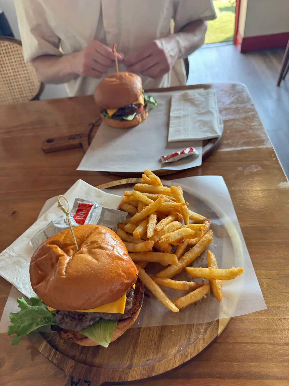 A close-up of a cheeseburger with lettuce and a side of golden french fries served on a wooden platter, with another burger and a person in the background.