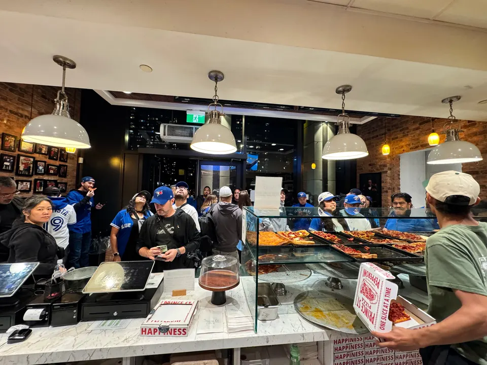 A busy counter at Prince Street Pizza in Wellington Place, with customers in line waiting for pizza slices displayed in a glass case, while a staff member in a green shirt holds a pizza box.