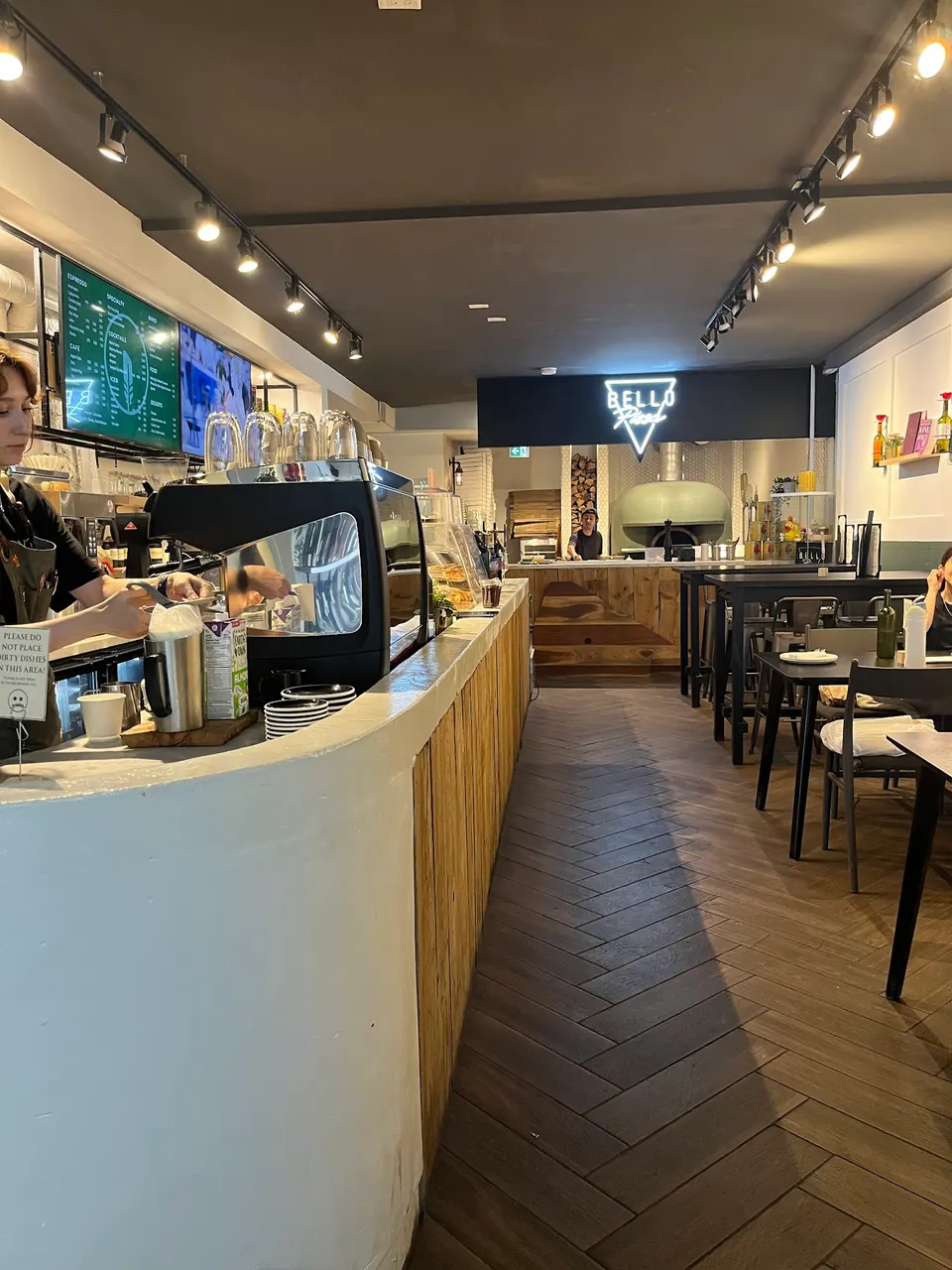 Interior view of Bello Pizza Toronto showing a long curved service counter on the left with espresso machine, a wooden herringbone floor, and dining tables on the right leading to a pizza oven in the back.