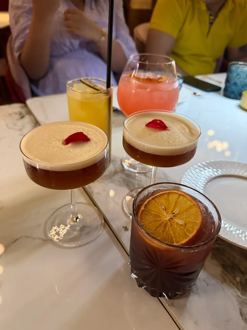 A selection of five colorful cocktails on a marble table at Aangan Toronto, including two espresso martinis with rose petals, a yellow drink with a lime wheel, a pink cocktail, and a dark drink with a dried orange slice.