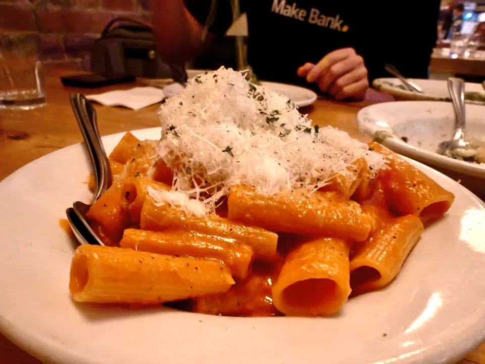 A plate of rigatoni pasta coated in a rich orange tomato sauce, topped with a generous mound of finely grated parmesan cheese and herbs, served on a wooden table.