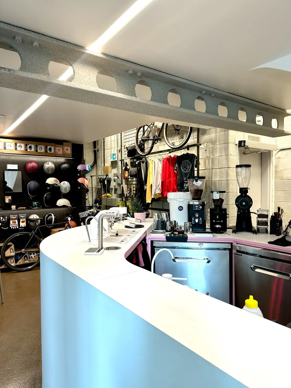 A modern curved white service counter at Fix Coffee + Bikes, featuring espresso equipment on one side and a display of bicycle helmets and accessories in the background.