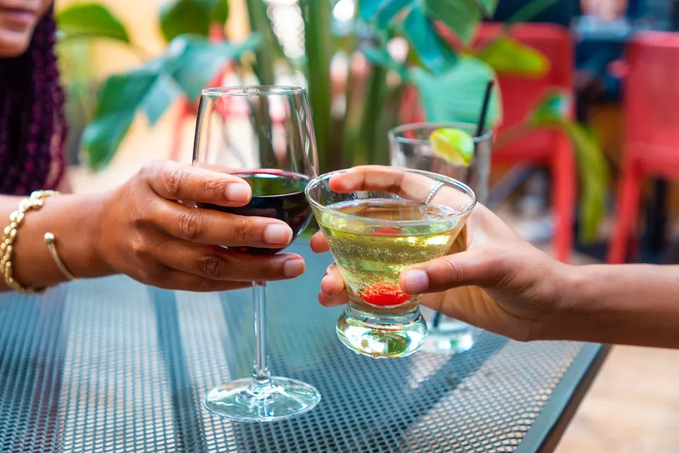 Two people clinking glasses in a toast over an outdoor patio table. One hand holds a wine glass filled with red wine, while the other holds a cocktail glass with a light-colored drink and a red berry garnish.
