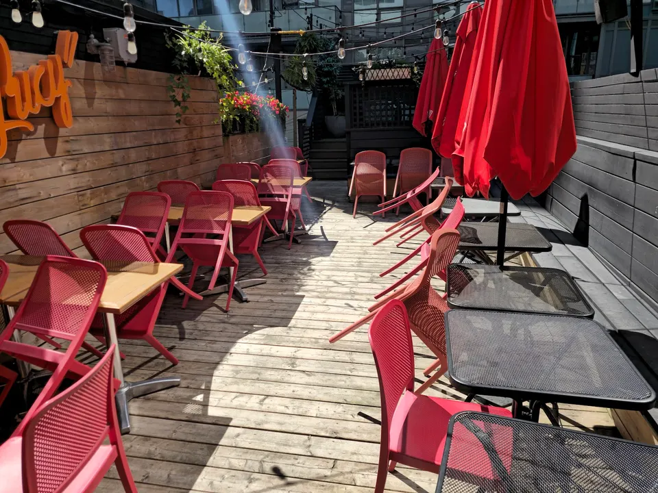 An outdoor wooden patio at Hey Lucy with rows of light wood tables paired with bright pink mesh chairs, and black metal tables with red umbrellas on the right side.
