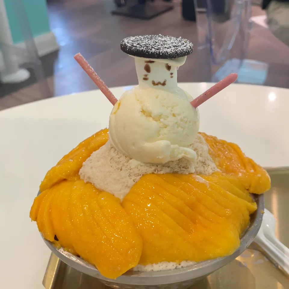 A bowl of mango shaved ice dessert topped with a snowman made of vanilla ice cream, a marshmallow head with a drawn face, an Oreo cookie hat, and pink biscuit stick arms.