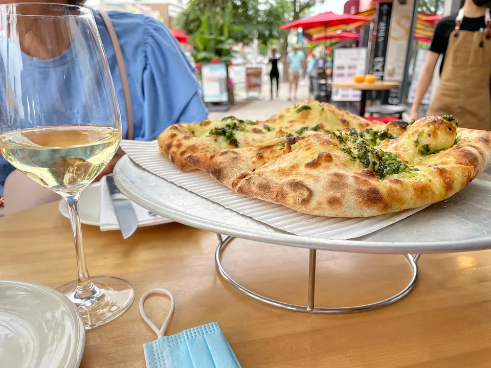 A freshly baked flatbread pizza with green herb topping sits on a raised metal stand next to a glass of white wine on an outdoor patio table.