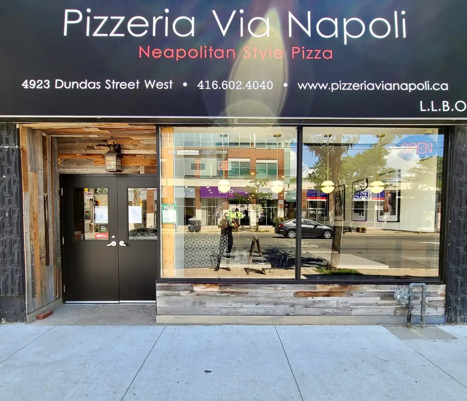 The storefront of Pizzeria Via Napoli featuring a black sign with white text, a double dark door entrance, and large glass windows showing reflections of the street.